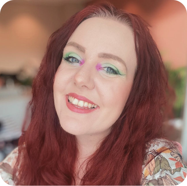 Arevya is a woman with mid-length dark red hair. She has a green-to-white gradient eyeshadow on her eyelids, fuchsia eyeshadow in the corner of her eyes, delicate liquid liner wings, and light red lipstick. She’s smiling widely at the camera.