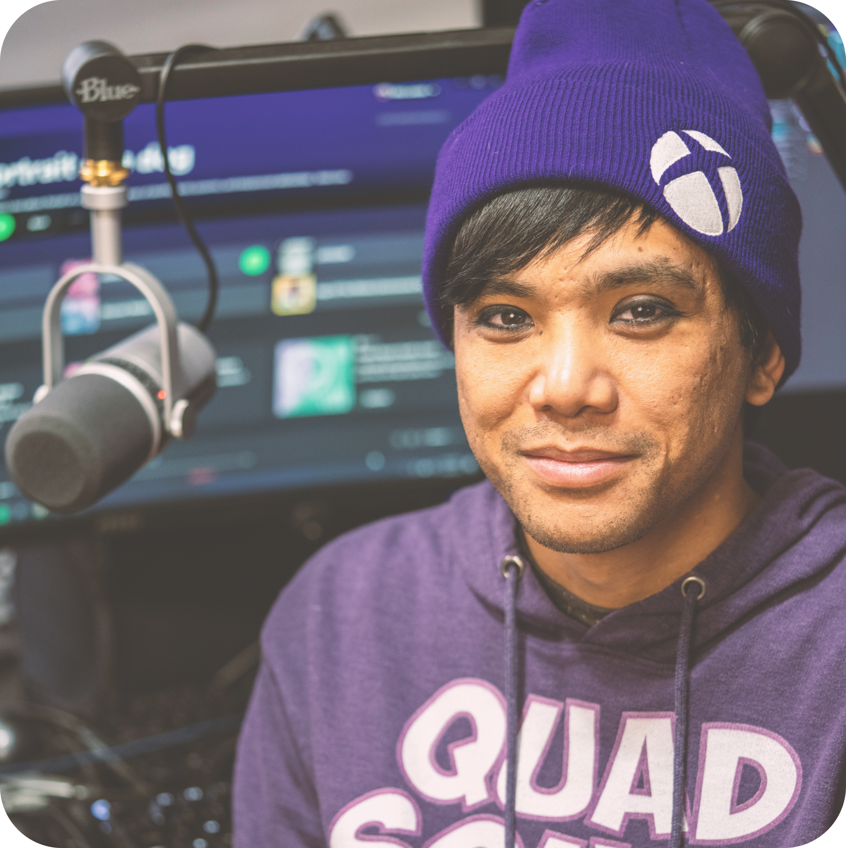 Mike is a man with brown eyes and short brown hair with sidebangs. He's wearing a purple tuque with the Xbox logo and a purple hoodie that reads Quad Squad. There's a microphone on a boom to his left, a television behind him, and he's smiling at the camera.