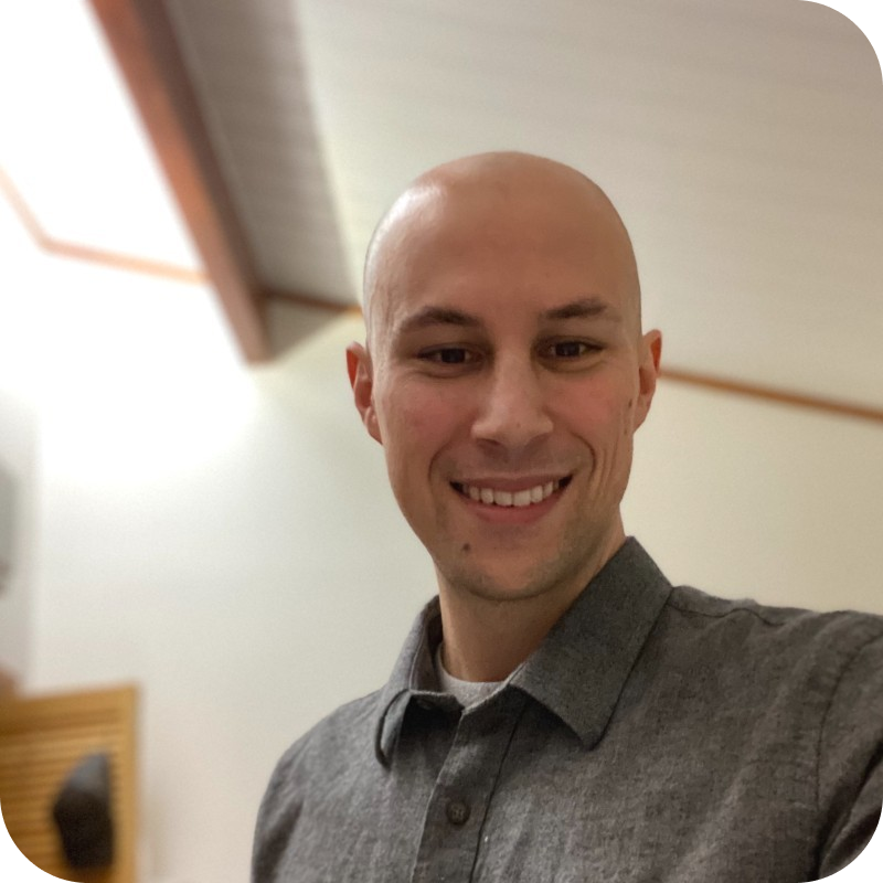 Noah Fishel is a man with brown eyes and a shaved head. He's wearing a grey button-up shirt and is smiling as he takes a selfie.