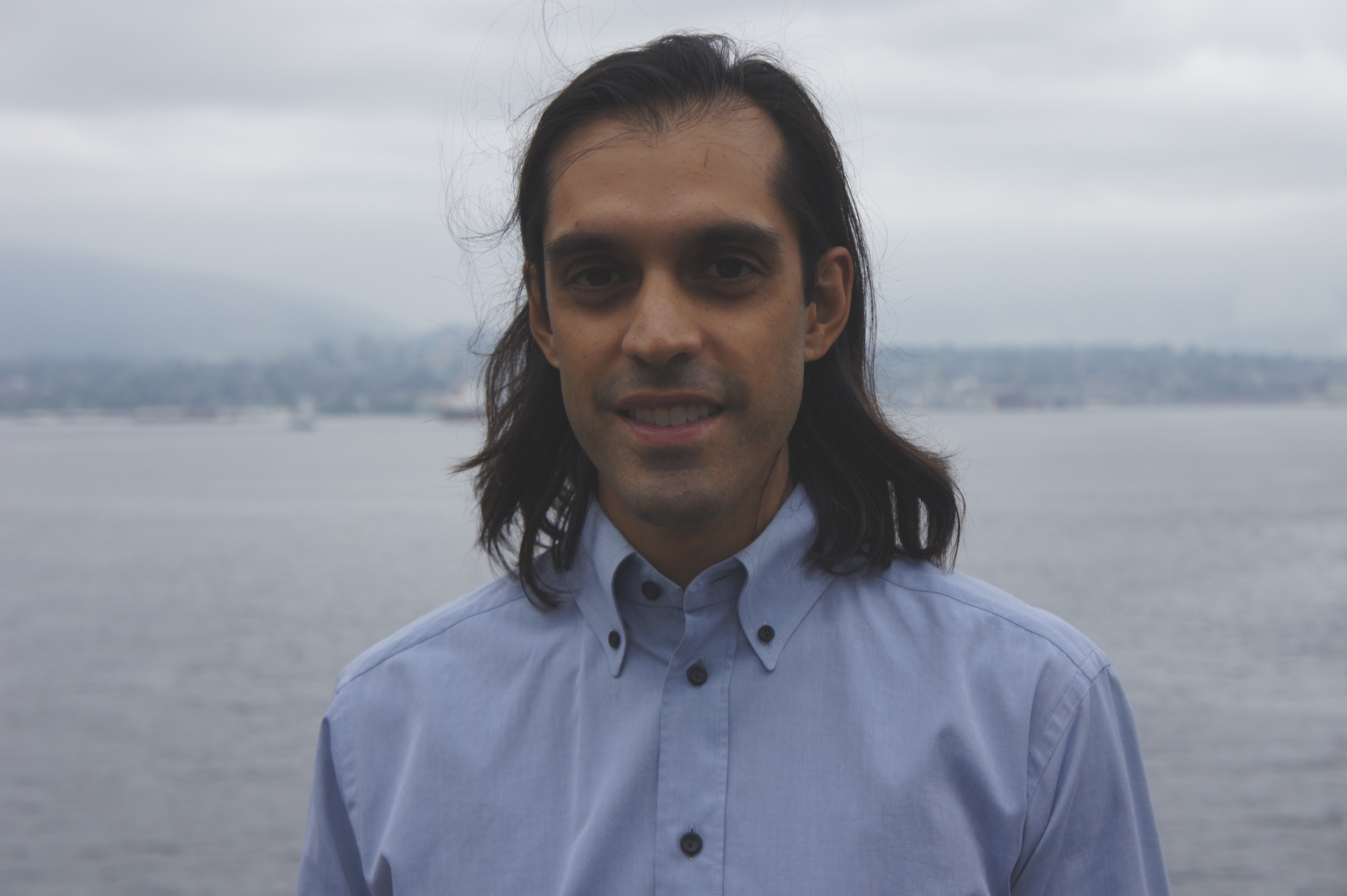 Noah is a 33-year old man with shoulder-length, dark brown hair and is wearing a blue button-down shirt. The Pacific Ocean is in the background.