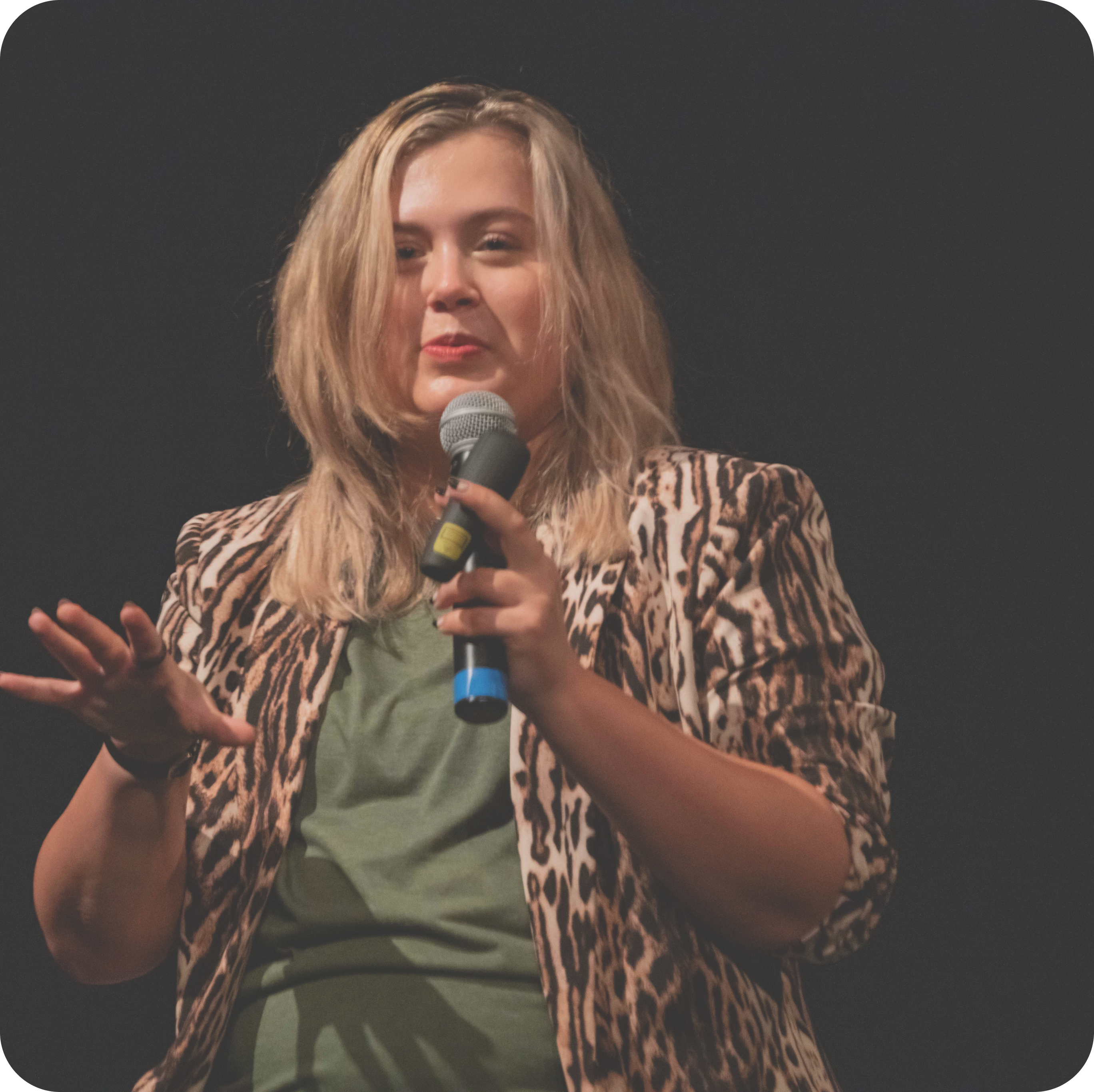 Rikki is a woman with medium length blond hair. She's wearing an olive green shirt, a leopard print blazer with the cuffs rolled up, and red lipstick. She's holding a microphone and is talking to the audience at a speaking event.